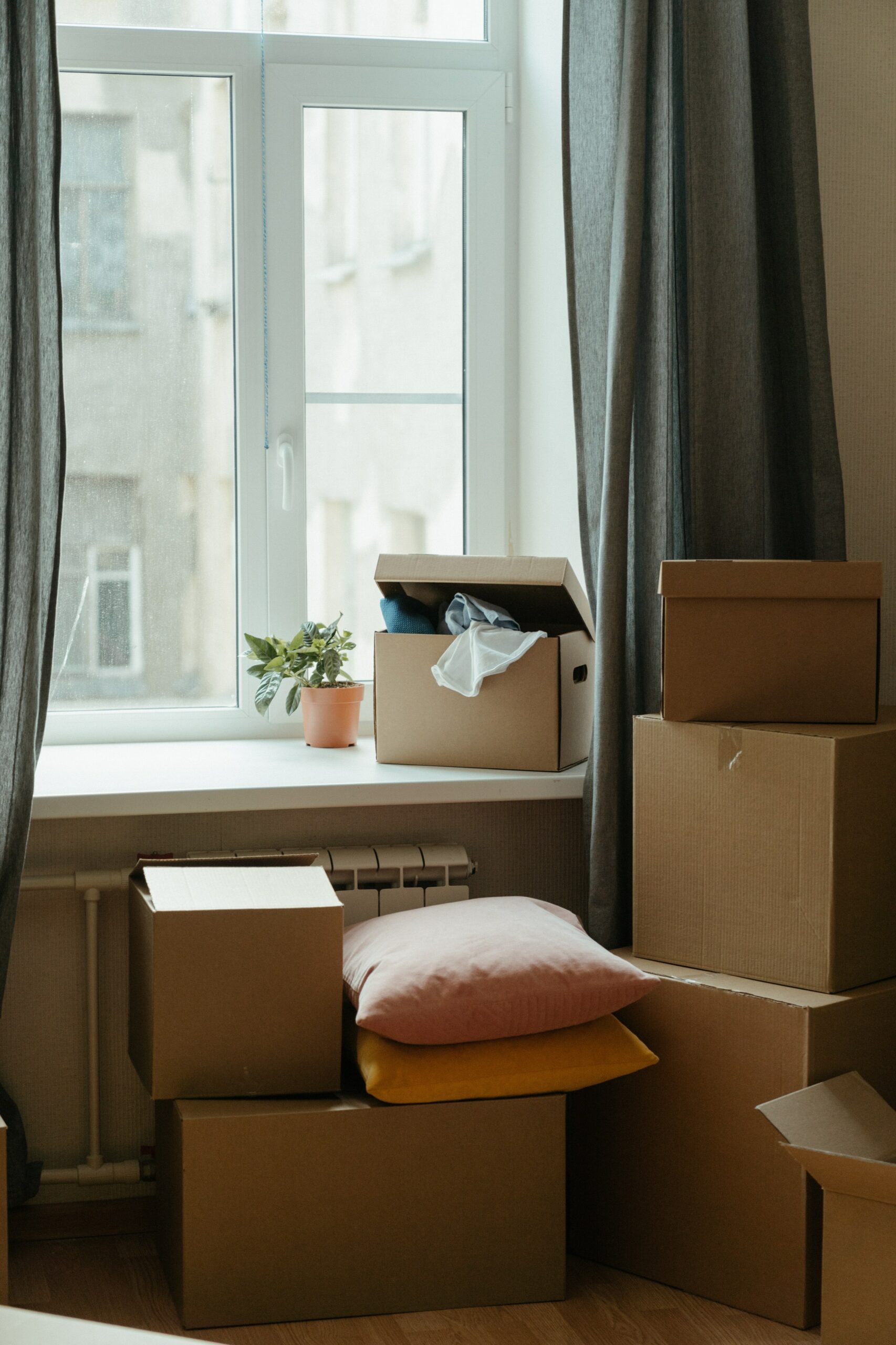 A window framed by curtains with several stacked cardboard boxes in front of it.