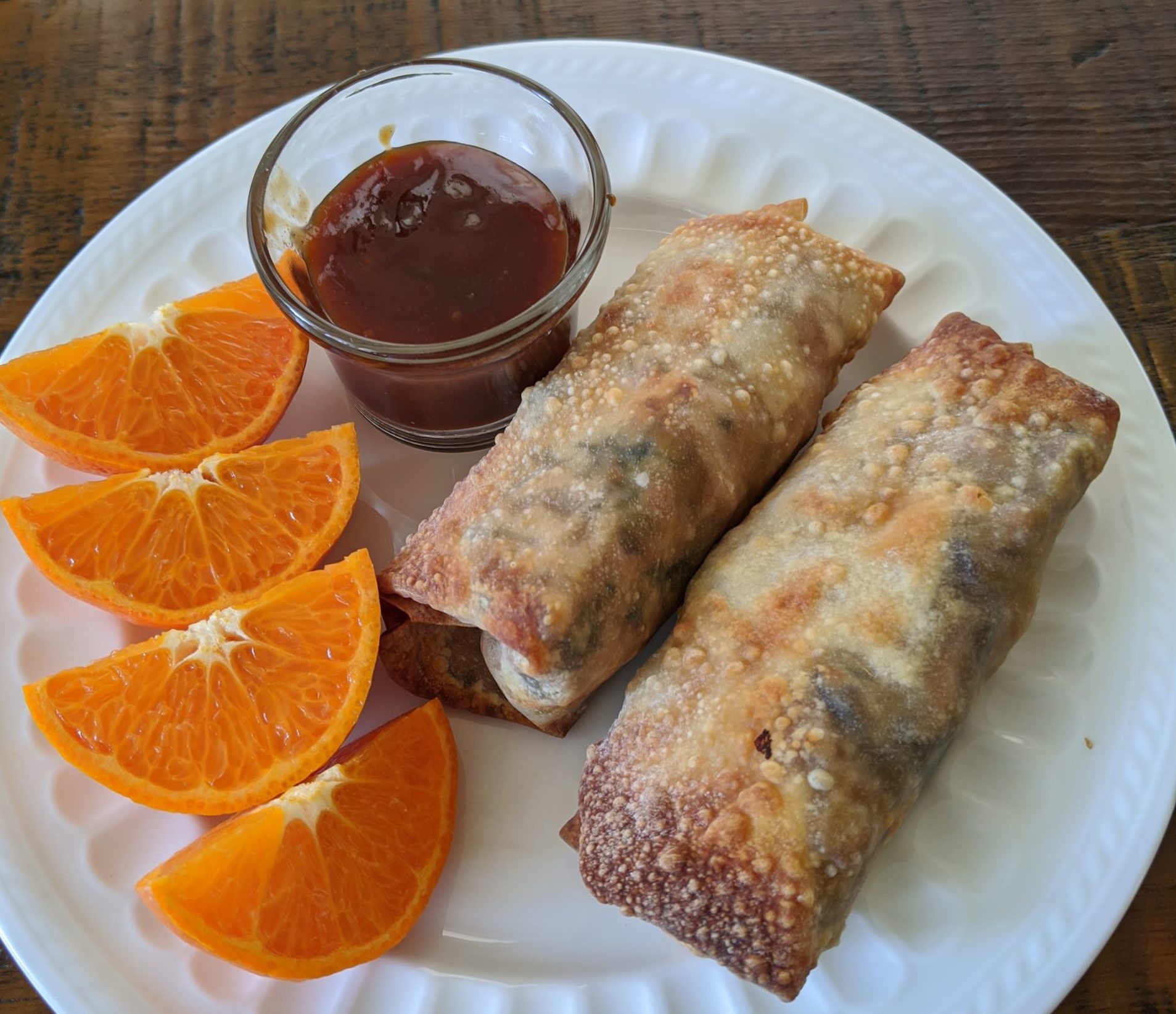 The Air Fryer Egg Rolls on a plate, accompanied by orange slices and a container of sauce.