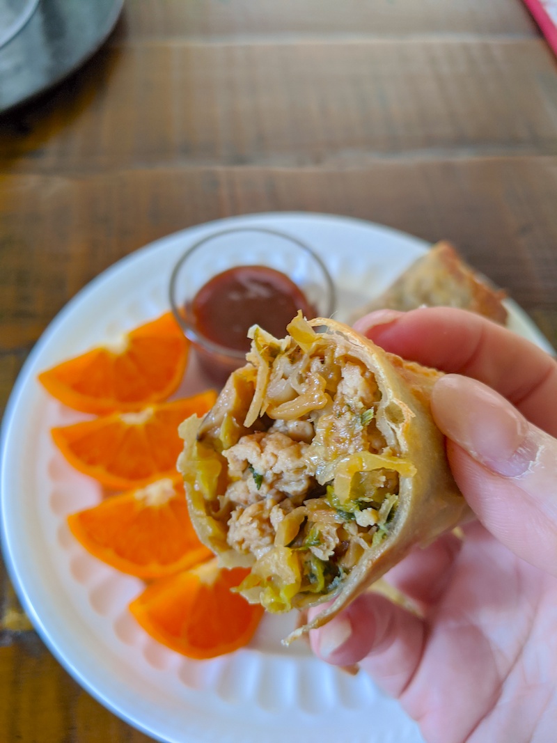 A half of the Air Fryer Egg Rolls being held up above a plate of orange slices.