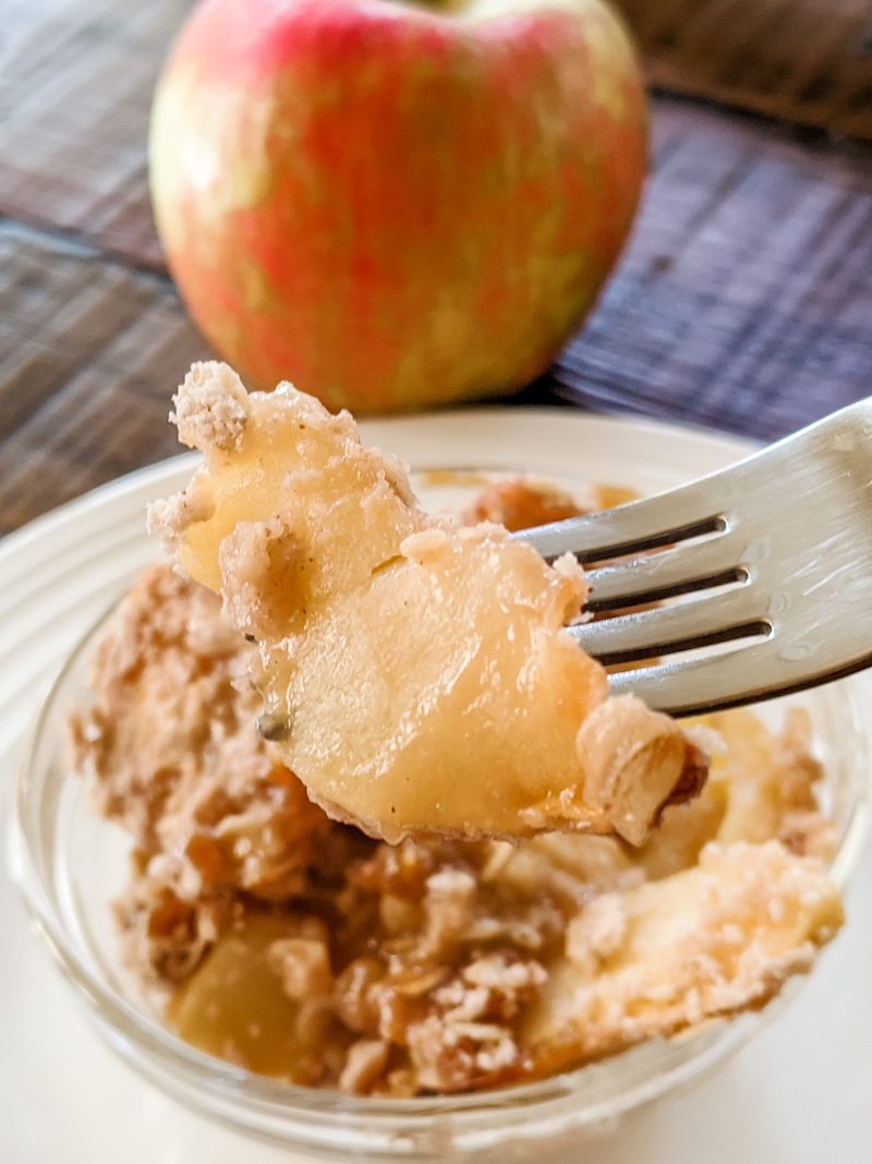 A fork holding up a piece of the finished Apple Crisp with an apple in the background