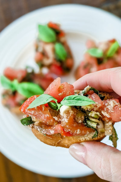 A hand holding a piece of the easy tomato bruschetta with more sitting on a plate in the background