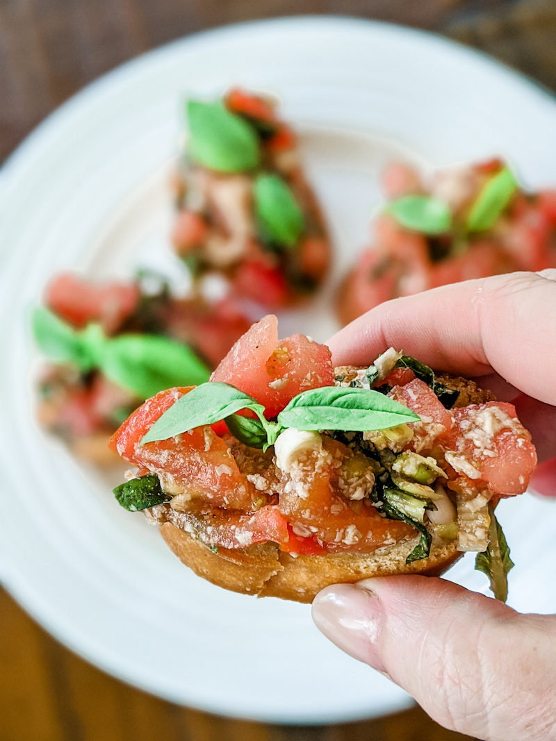 A hand holding a piece of the easy tomato bruschetta with more sitting on a plate in the background