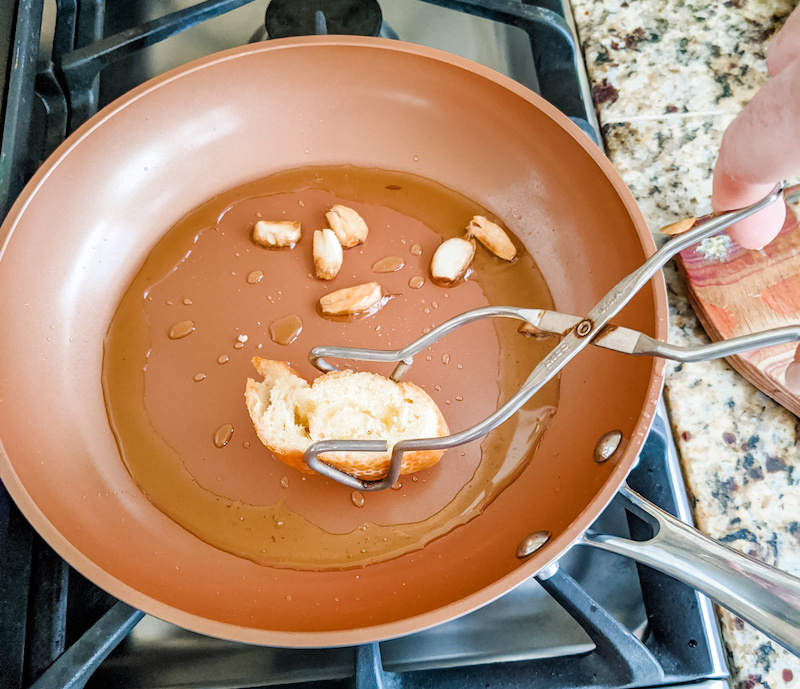 Tongs flipping a piece of the baguette in the garlic and olive in a pan