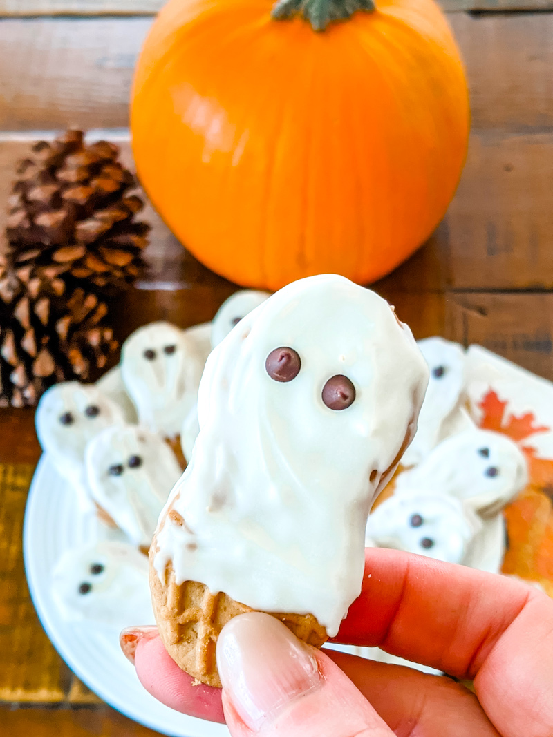 A finished Easy Nutter Butter Ghosts being held above a plate of other ones, as well as a pumpkin and pinecone.