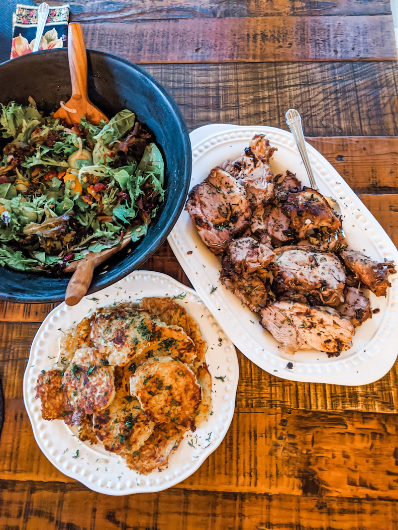 The Boston Pork Butt Roast on a platter along with a green salad, potatoe pancakes - all on a wooden table.