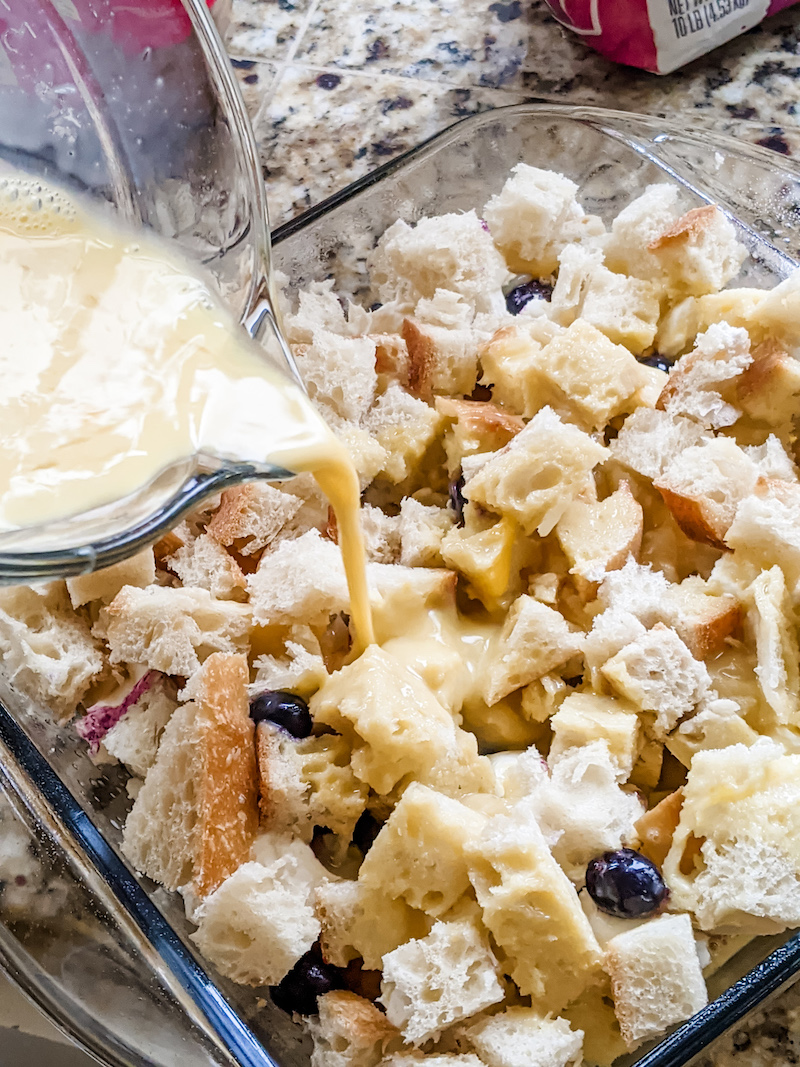 The liquid ingredients being poured into the casserole dish filled with bread and blueberries.