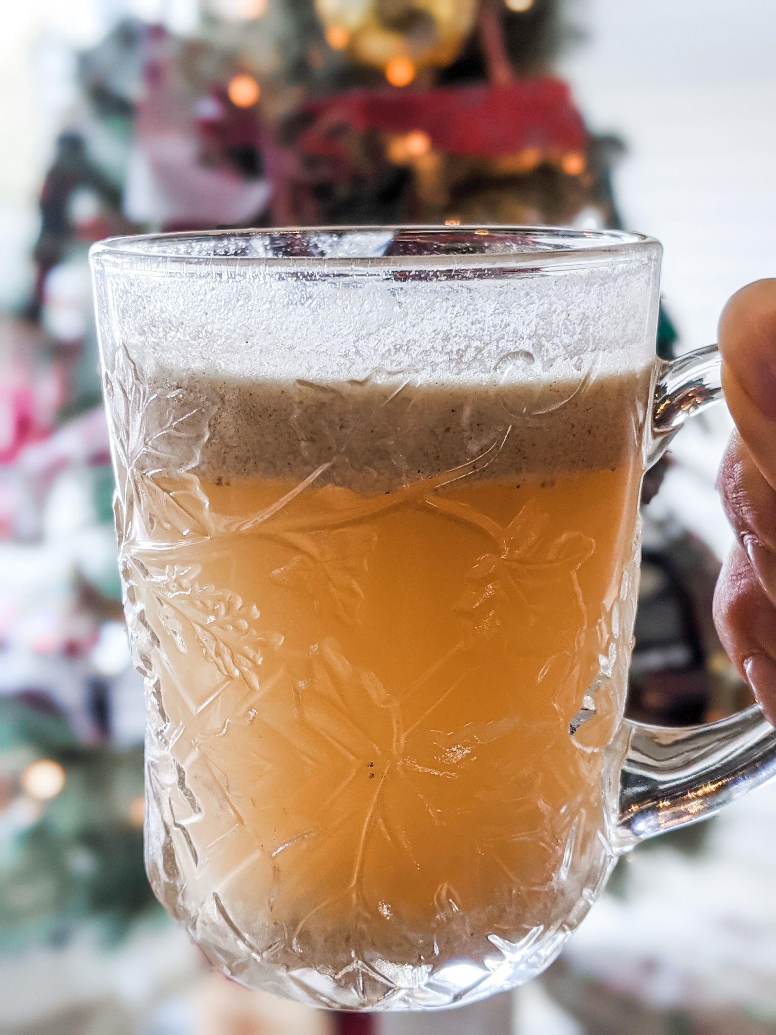 Marie's Easy Hot Buttered Rum in a clear mug, held in front of a Christmas Tree