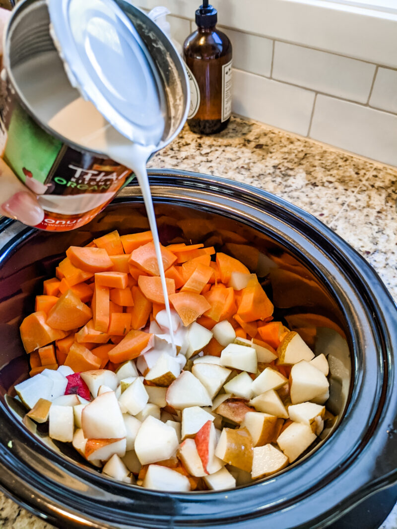 A hand pouring in the coconut milk into a slow cooker with cubed carrots and pears