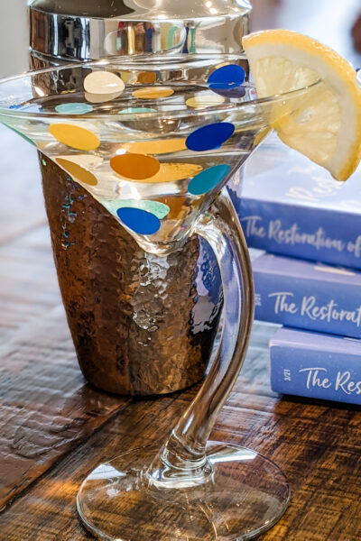 The finished DIY Polka Dot Martini Glasses with a lemon wedge, in front of The Restoration of Celia Fairchild paperbacks