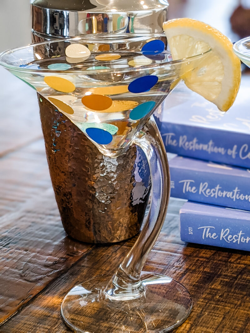 The finished DIY Polka Dot Martini Glasses with a lemon wedge, in front of The Restoration of Celia Fairchild paperbacks