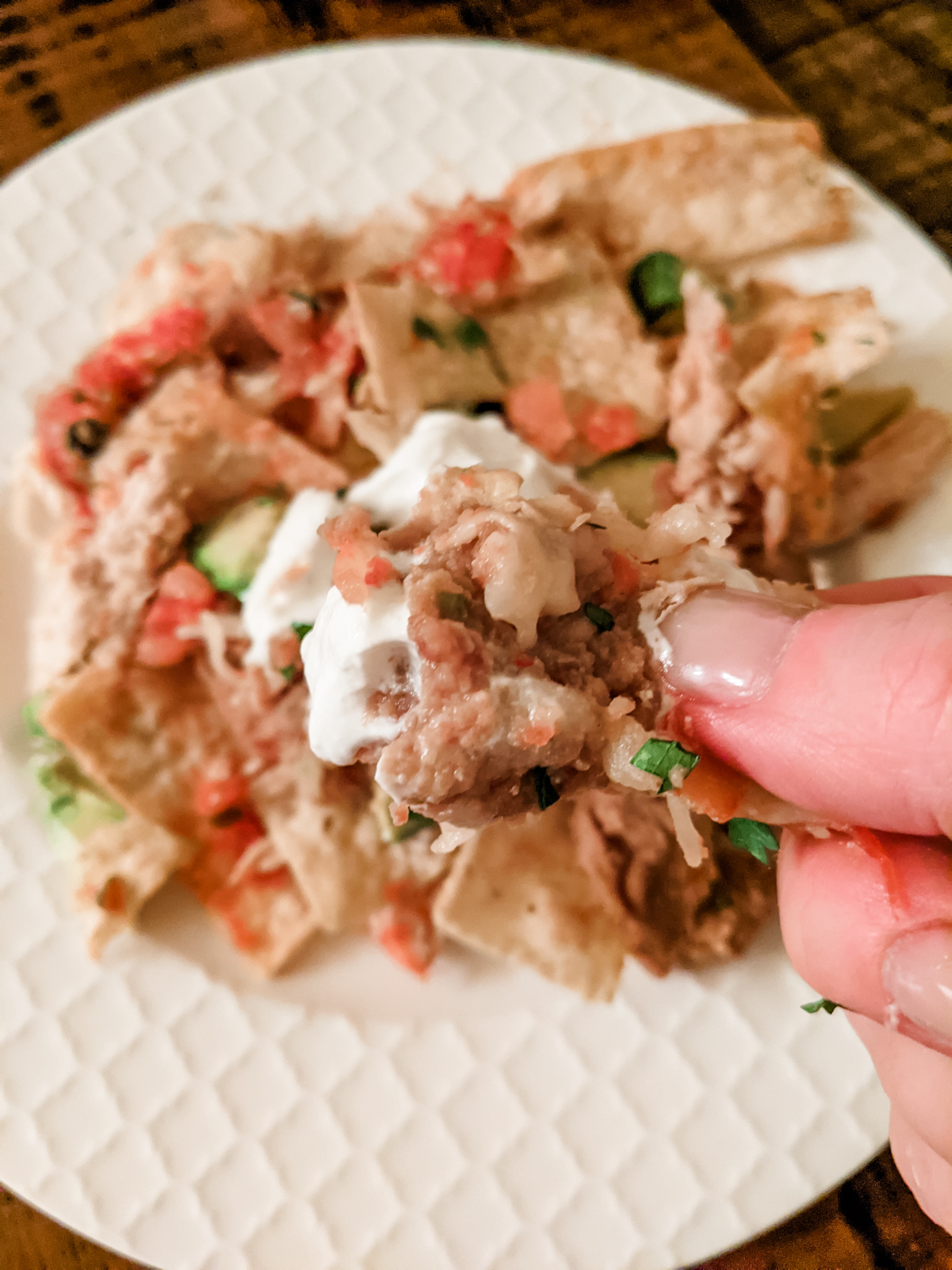 A hand holding a piece of the Healthier Chicken Nachos above a plate of them.