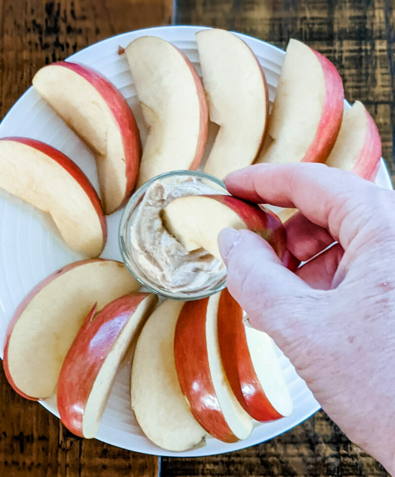 Apple slices spiraled out on a plate, with a hand dipping one into a clear container of dip - a healthy snack from home.