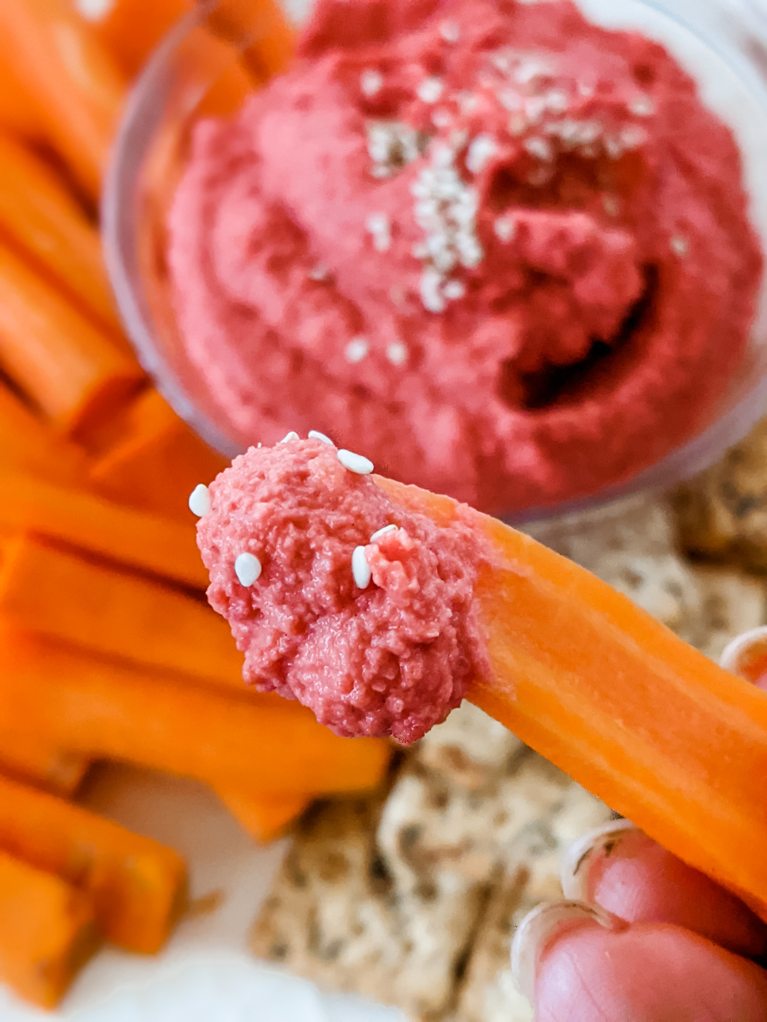 A carrot with a dab of the Homemade Beet Hummus, held above more of the hummus and carrots.