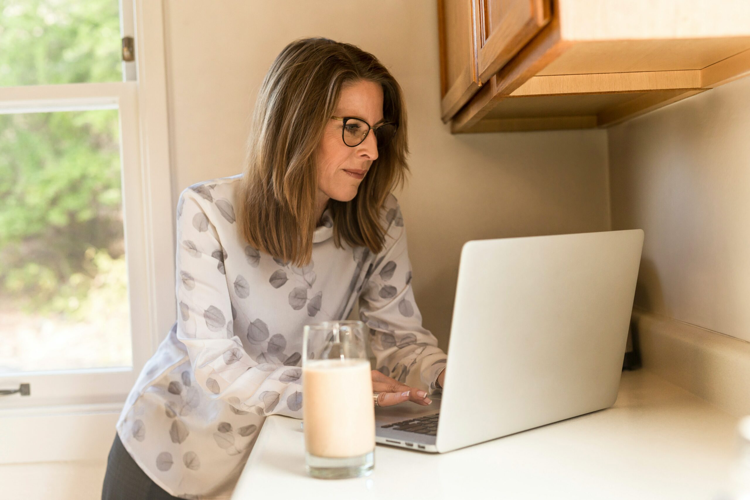 A woman in a kitchen wearing glasses looks at her laptop