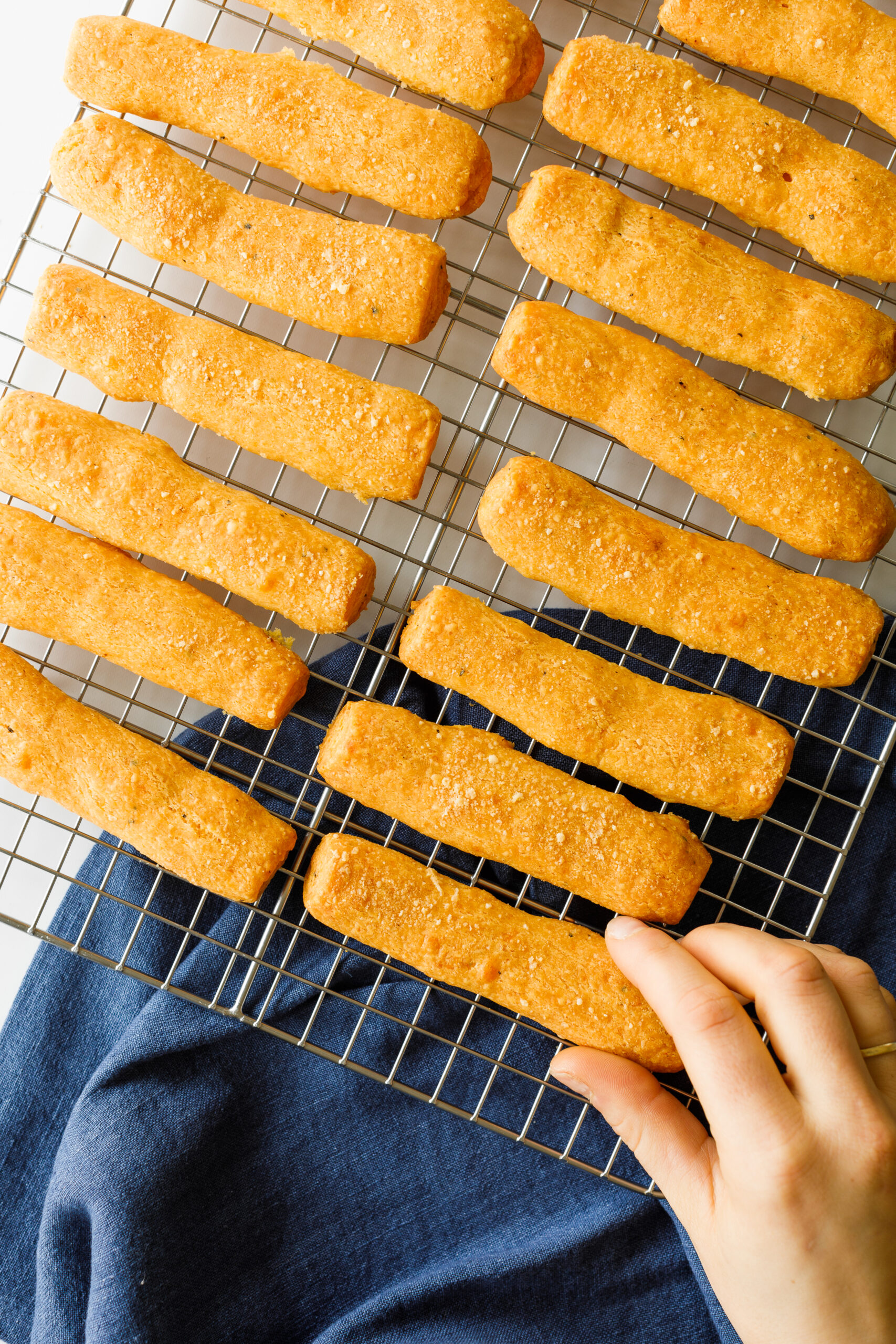 The Parmesan and Sharp Cheddar Cheese Straws on a cooling rack, with a hand reaching for one.