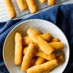 Marie's Parmesan and Sharp Cheddar Cheese Straws on a cooling rack and a bowl, with a blue tablecloth underneath