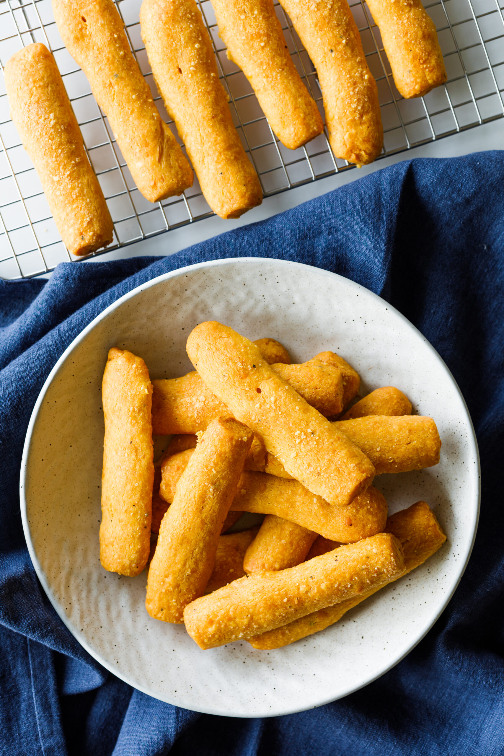 Marie's Parmesan and Sharp Cheddar Cheese Straws on a cooling rack and a bowl, with a blue tablecloth underneath