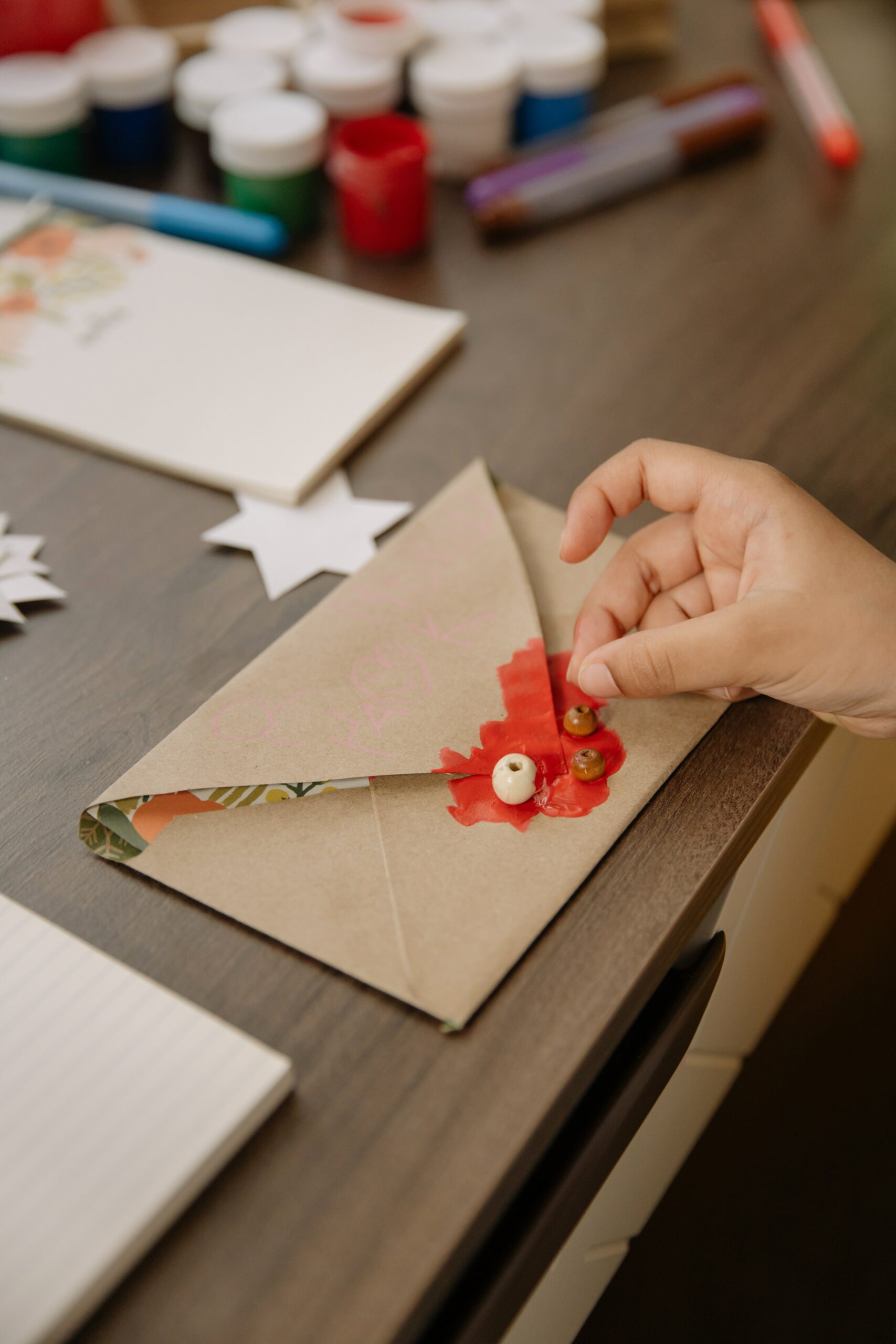 A small hand glues a bead on to a hand-decorated brown envelope, with crafting supplies in the background - a great way of keeping in touch with kids 