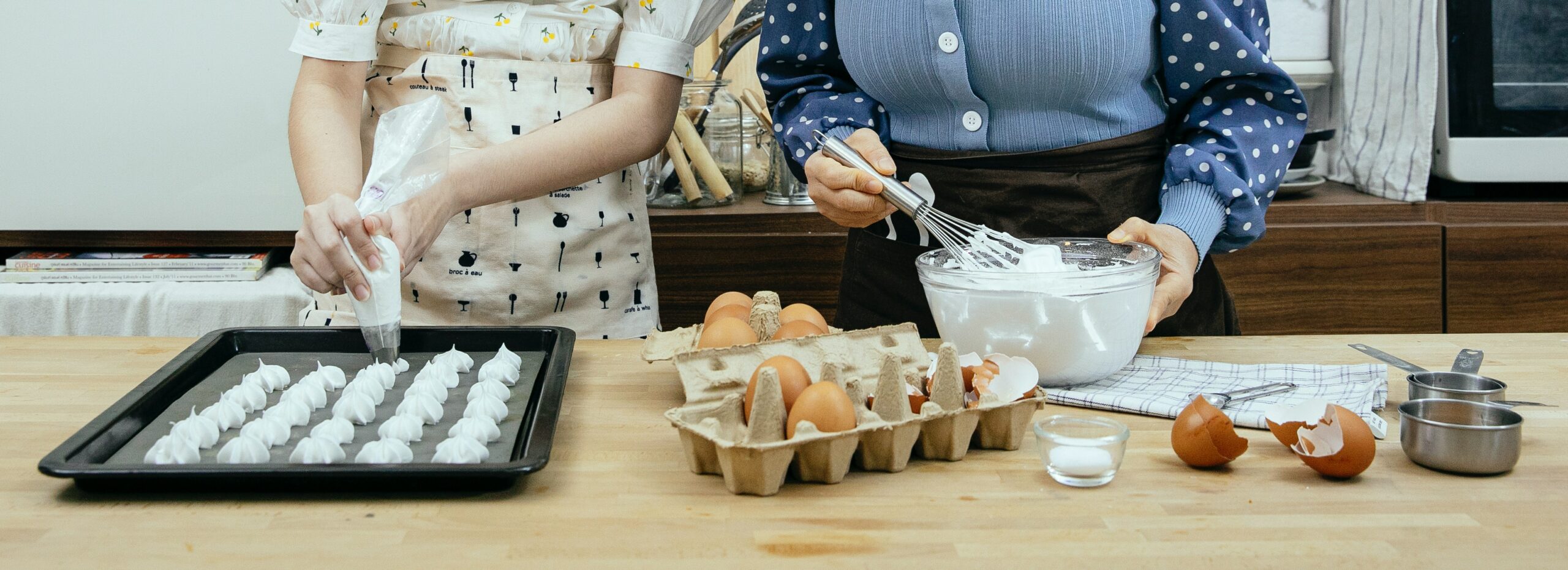 Two women in aprons prepare a baking pan, surrounded by ingredients such as eggs