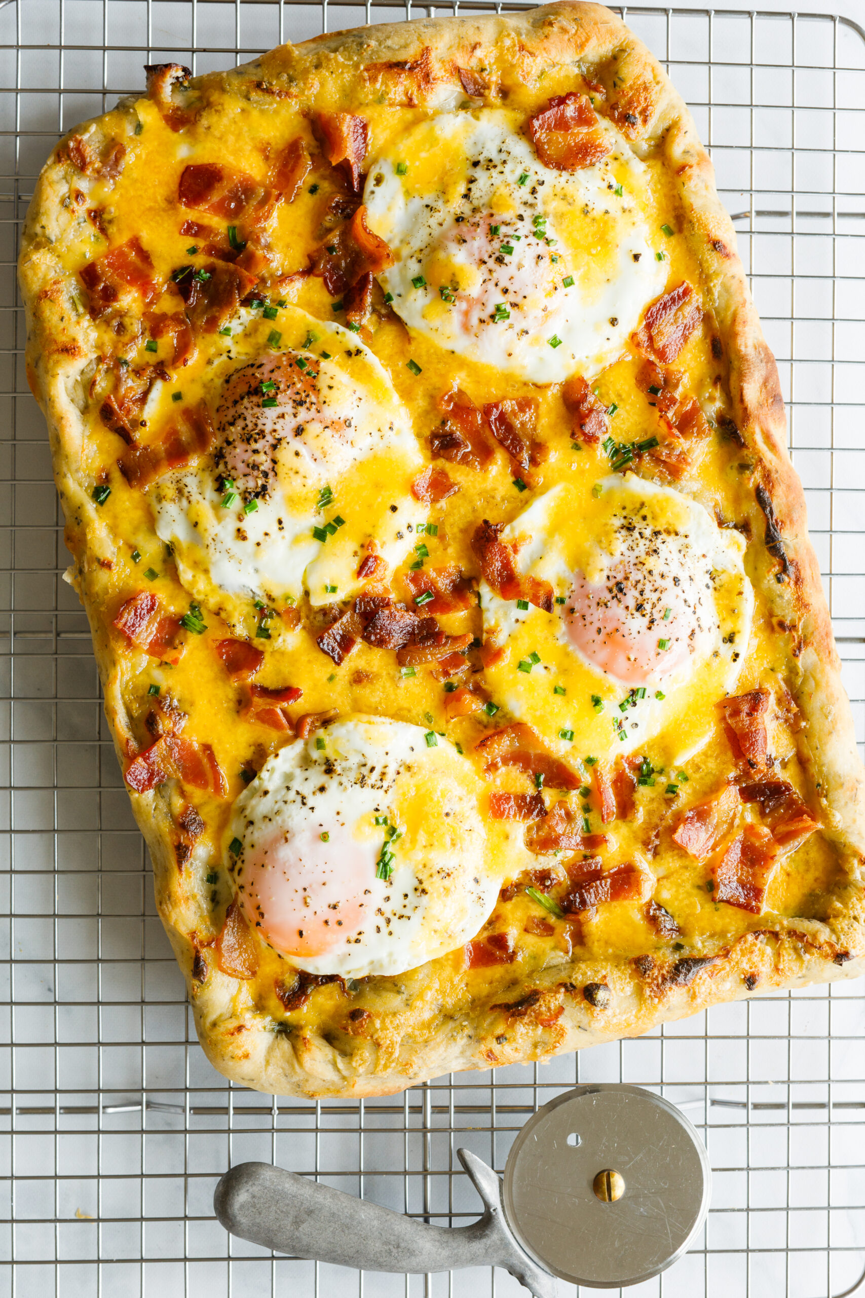 The Easy Bacon and Egg Pizza on a cooling rack with a pizza cutter