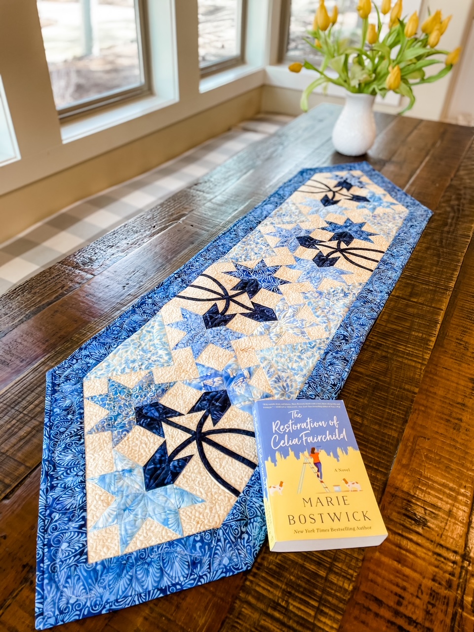 The quilted table runner on a wooden table, with Marie's novel and vase of flowers on top of it. 