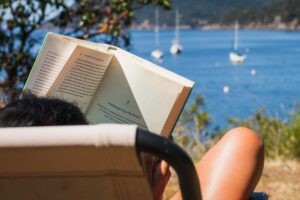 A woman reading a book in front of a marina - summer 2021 reading list