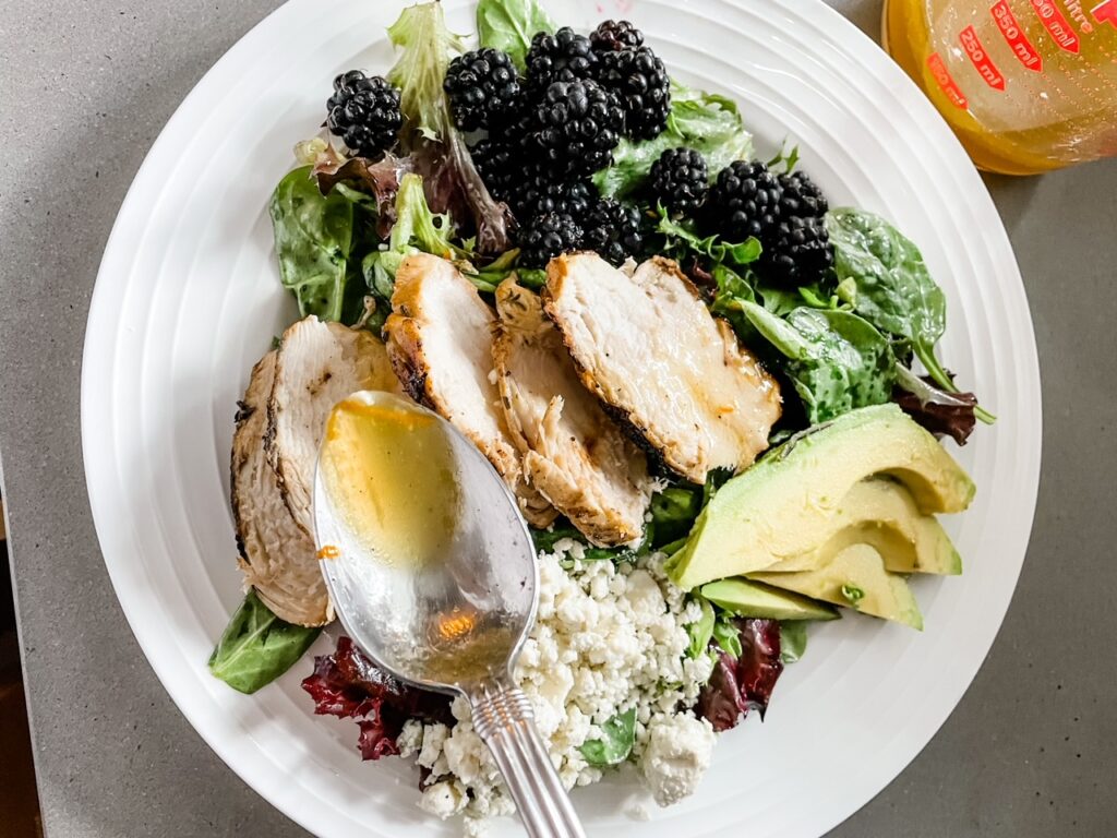 A white plate filled with the Blackberry and Grilled Rosemary Chicken Salad and a spoon above drizzling extra vinaigrette on top