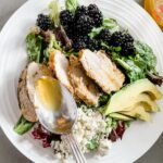 A white plate filled with the Blackberry and Grilled Rosemary Chicken Salad and a spoon above drizzling extra vinaigrette on top