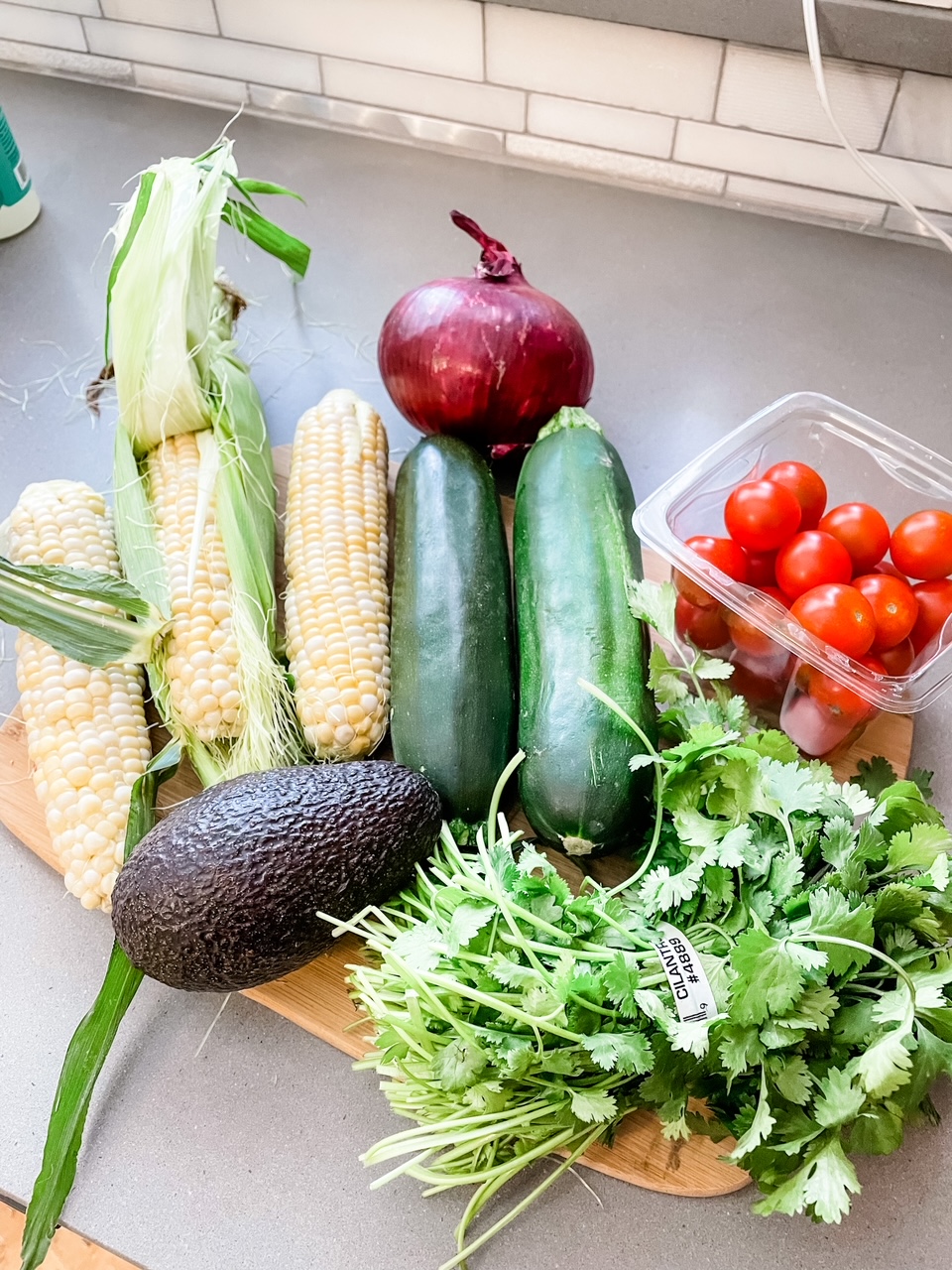 Corn, an avocado, cherry tomatoes, cucmbers, onions, and herbs on a wooden cutting tray