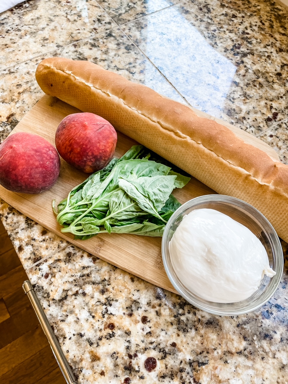 The ingredients for the Fresh Peach and Burrata Bruschetta - a loaf of bread, two peaches, basil leaves, and burrata, on a wooden cutting board
