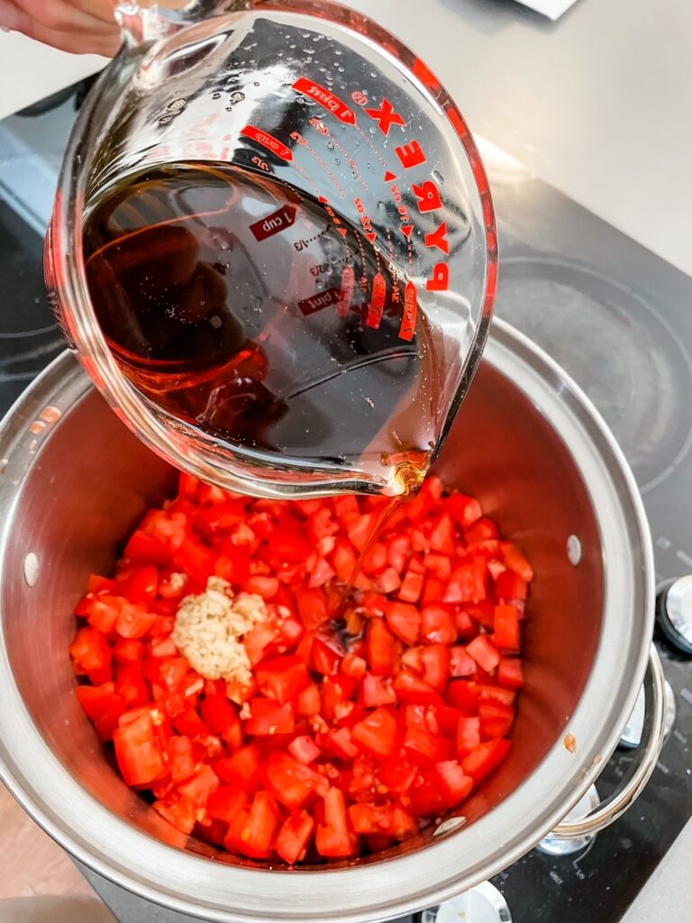 A measuring cup pouring maple syrup into the pot filled with tomatoes for the Easy Tomato Jam with Maple