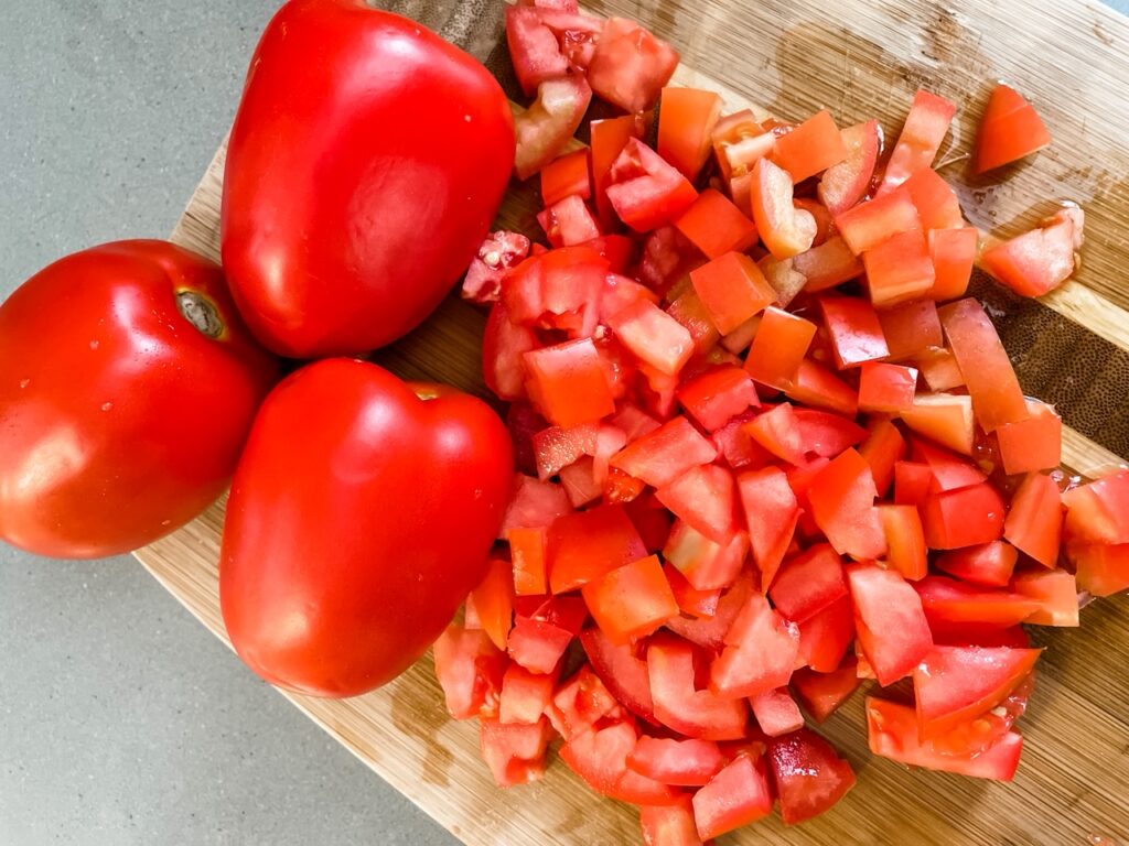 Some whole roma tomatoes alongside the chopped ones on a wooden cutting board