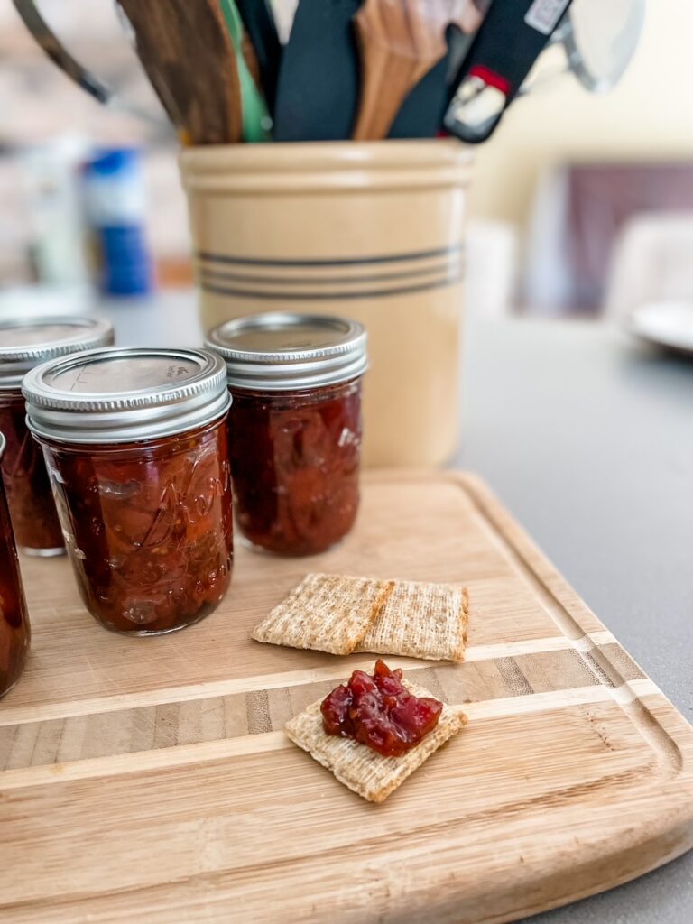 A cracker spread with Easy Tomato Jam with Maple in front of the clear jars of the jam