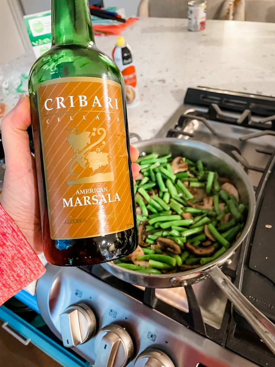 Marie holding a bottle of marsala overr a pan cooking greenbeans and mushrooms