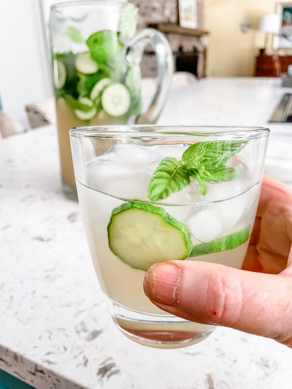 Marie's hand holding a Coconut-Lime Cucumber Mocktails in front of the pitcher