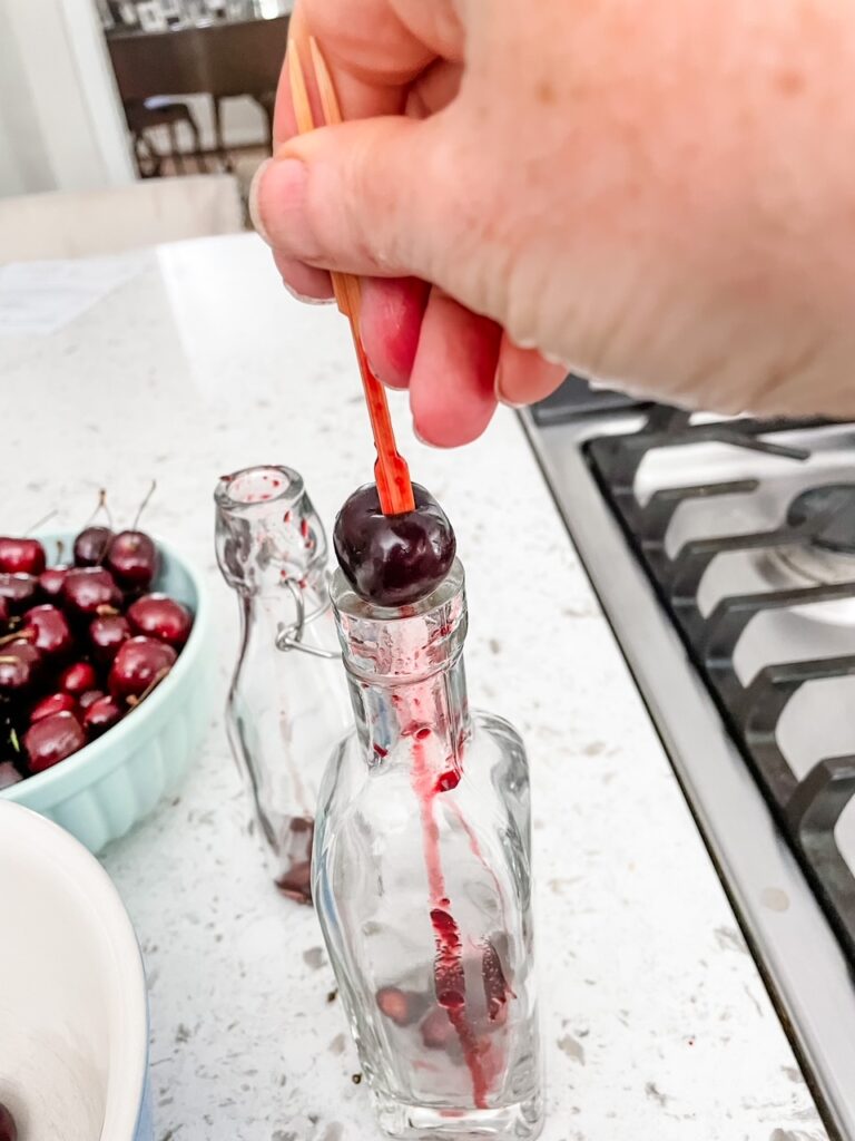 A cherry being pitted by having it placed in a bottle and pushed on by a flat utensil as part of how Make Your Own Cherry Liqueur 