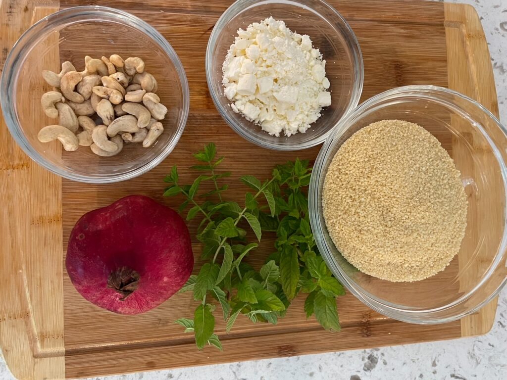 The ingredients for the Couscous Recipe laid out on a wooden cutting board - almonds, feta, couscous, mint, and pomegranate 