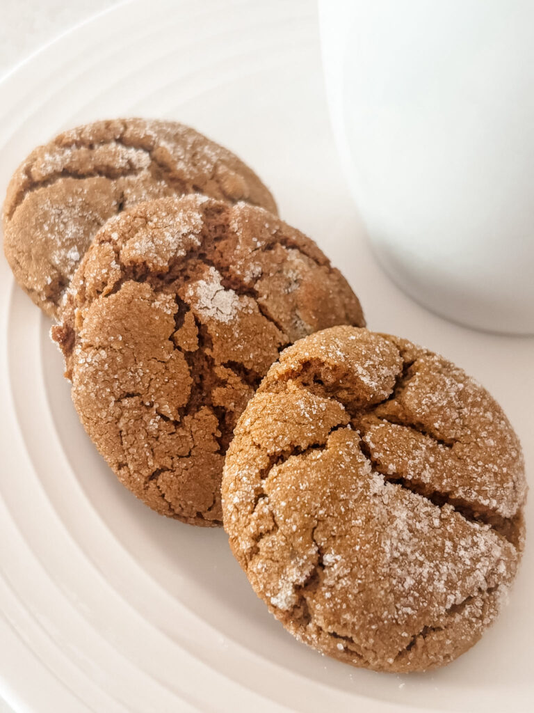 Three chewy triple ginger molasses cookies stacked on a white plate next to a white mug, dusted with sparkling sugar