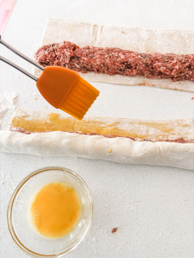 The pastry with the sausage mixture, being sealed with an egg wash