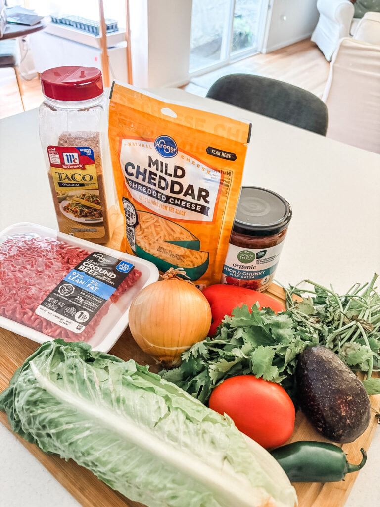 Ingredients for a low-carb taco board laid out on a counter, including ground beef, McCormick taco seasoning, romaine lettuce, avocado, roma tomatoes, jalapeño, shredded cheese, salsa, and fresh cilantro
