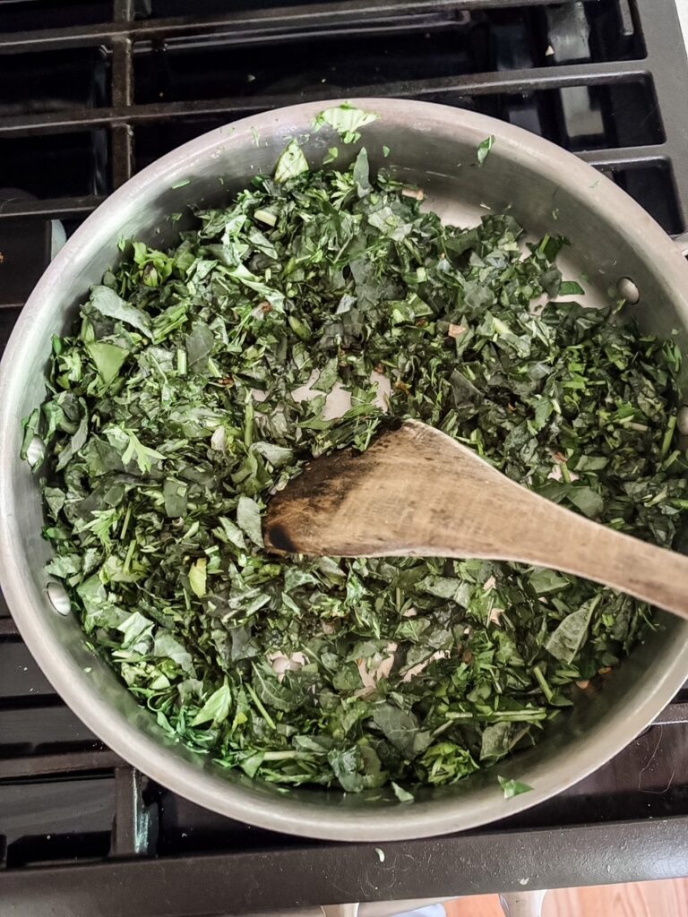 Chopped kale, carrot tops, and basil wilting in a heavy bottomed saucepan with shallots and garlic