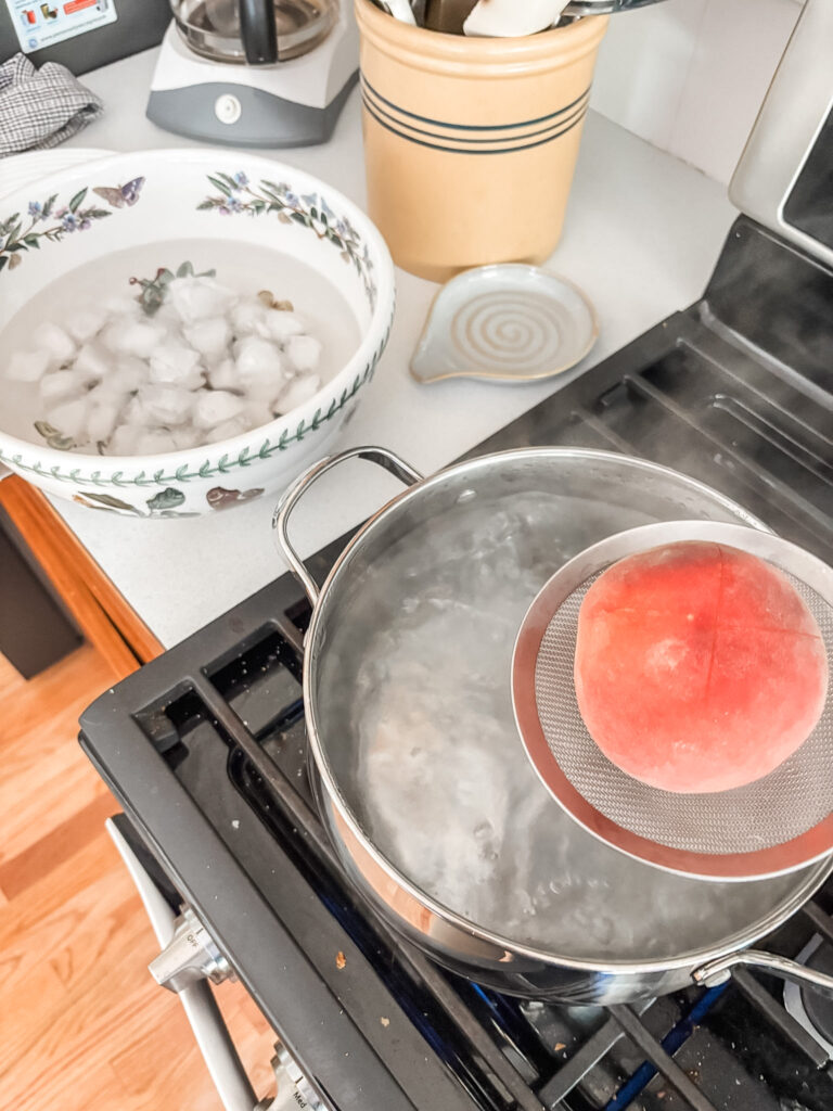 Peaches being lifted from boiling water with a slotted spoon to plunge into an ice water bath