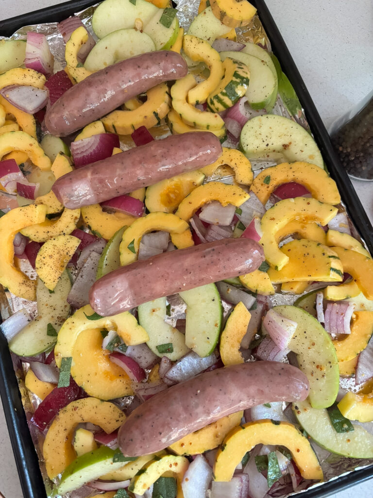 Glazed squash mixture spread on a foil-lined baking sheet with whole chicken apple sausages on top, ready for the oven